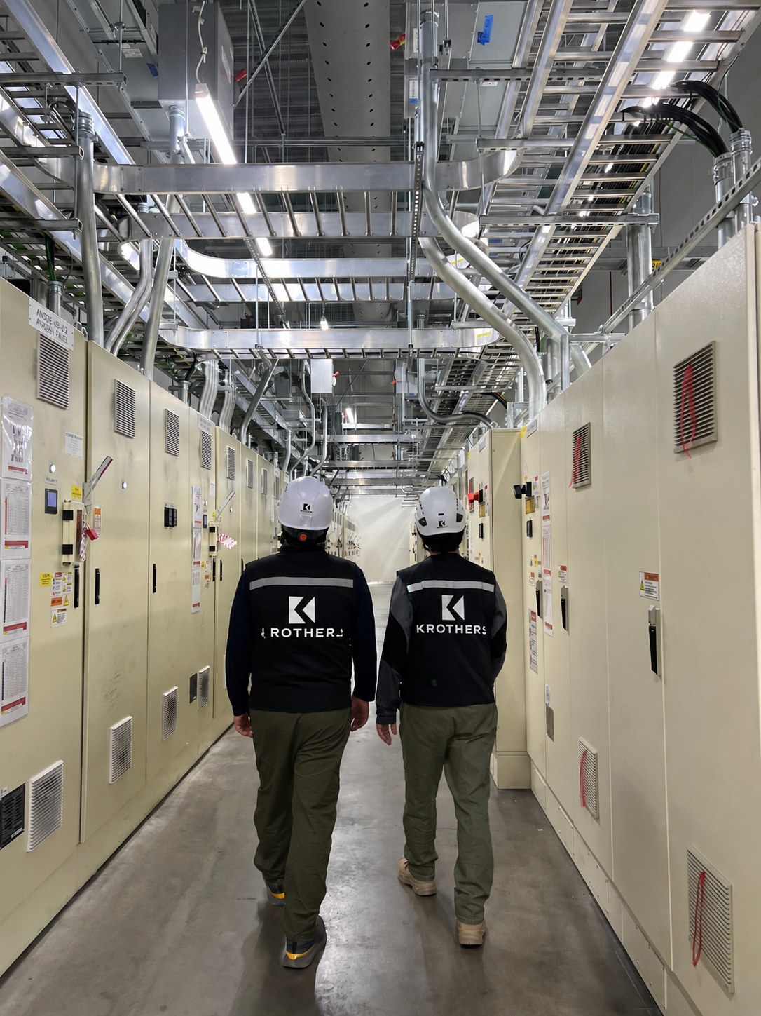 Two KROTHERS crew members in branded vests walking a corridor of installed electrical switchgear panels.