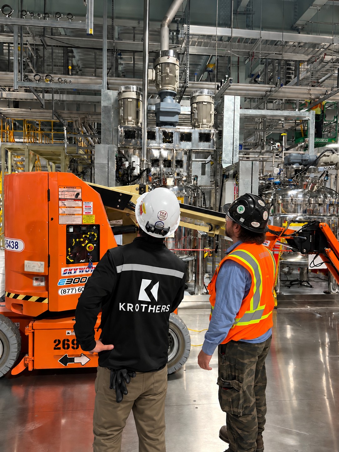 A KROTHERS crew member in branded workwear and a technician in a high-visibility vest review an orange boom lift beside stainless process equipment.