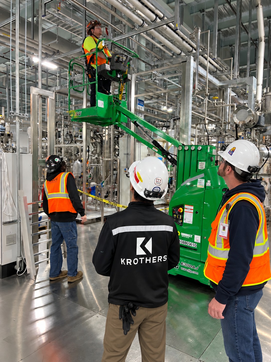 A KROTHERS crew watches a colleague elevated in a boom lift basket inspecting overhead piping in a stainless process facility.