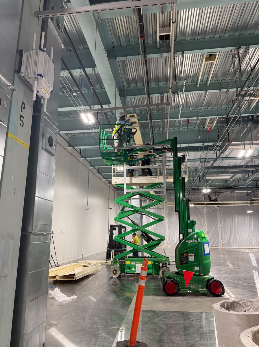 A field technician works from the guarded platform of a lift beside overhead industrial supports.