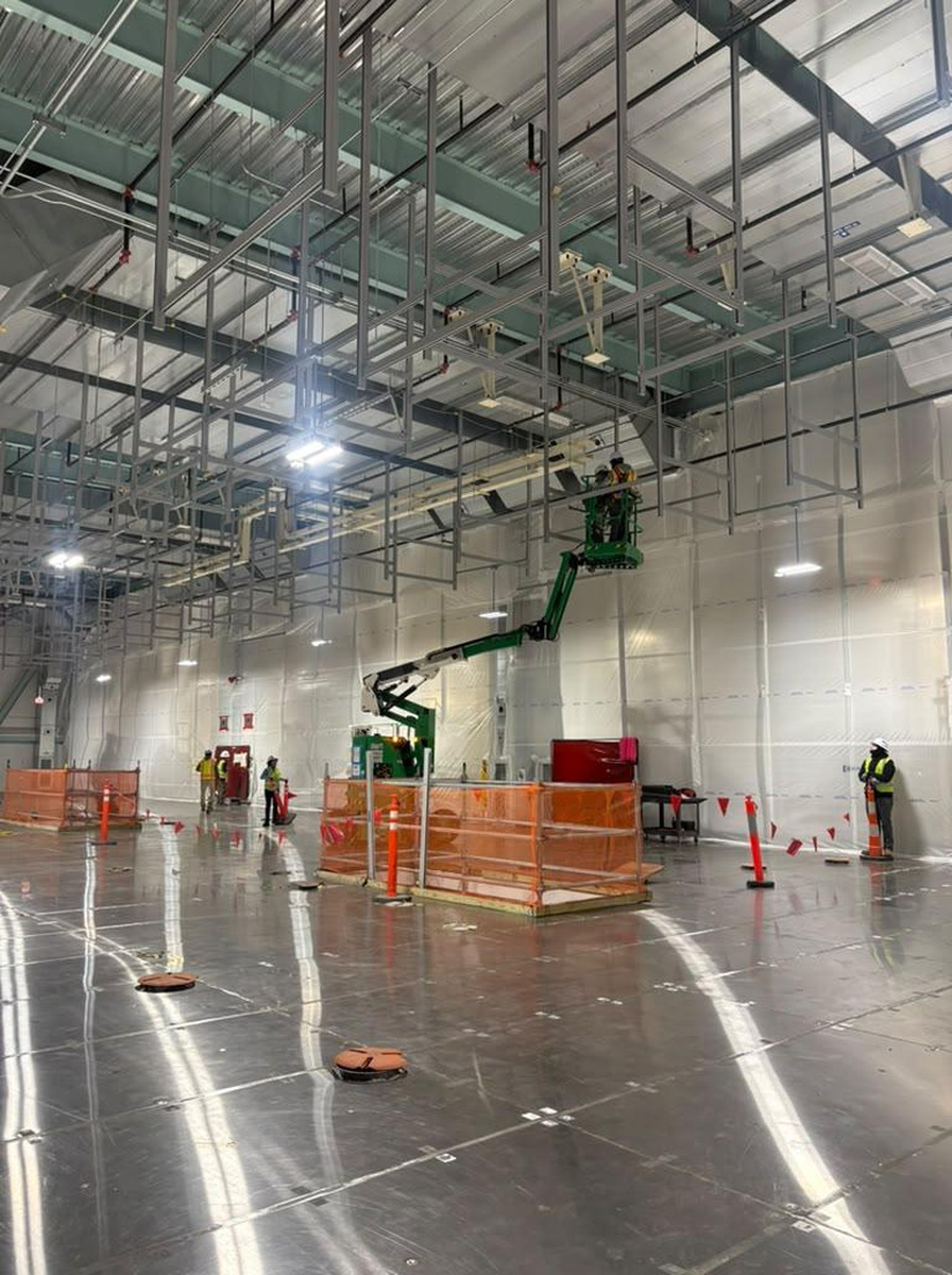Elevated installation work takes place in a barricaded industrial hall with a ground observer outside the work zone.
